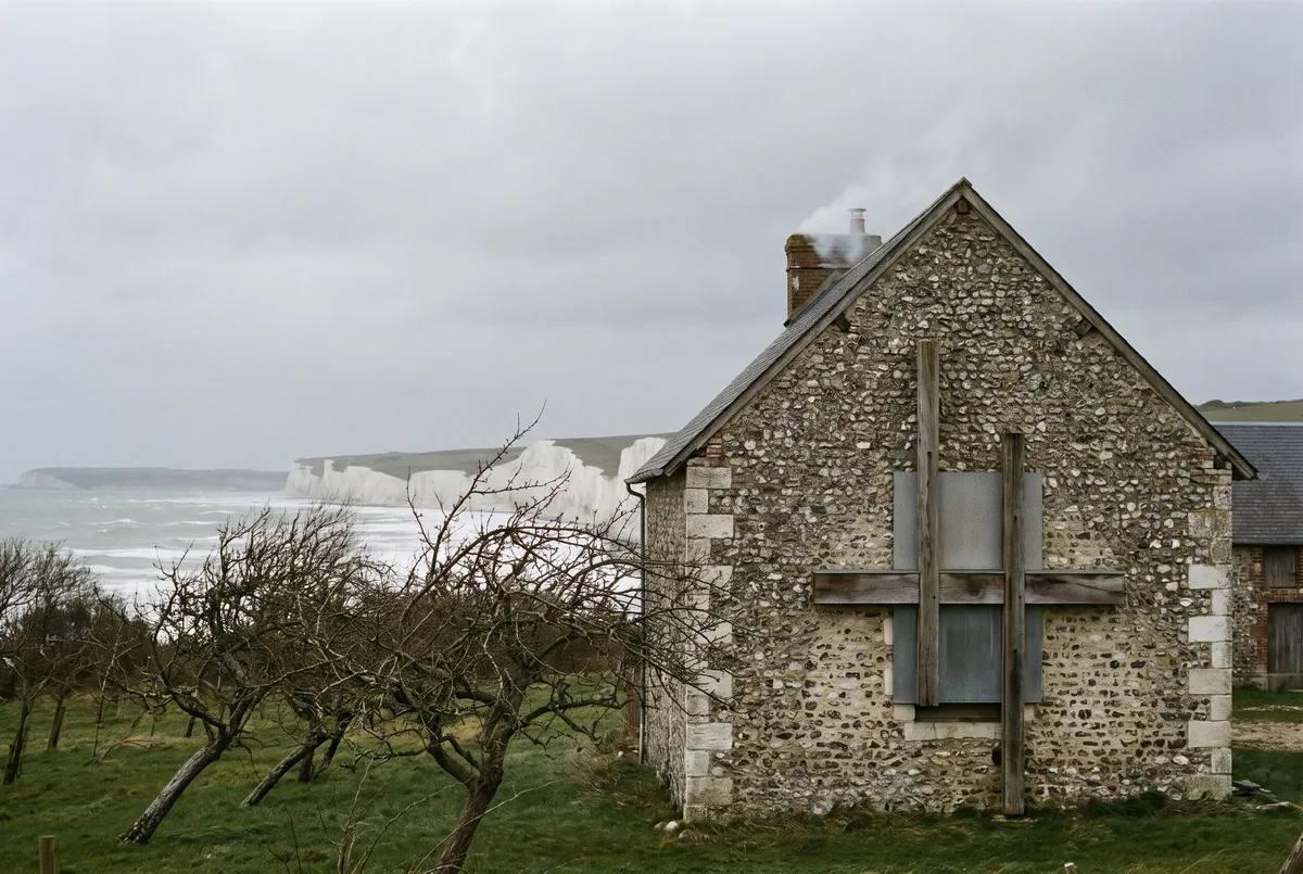 A stone barn near Fécamp under a grey sky, a dead apple orchard in front and chalk cliffs in the distance.